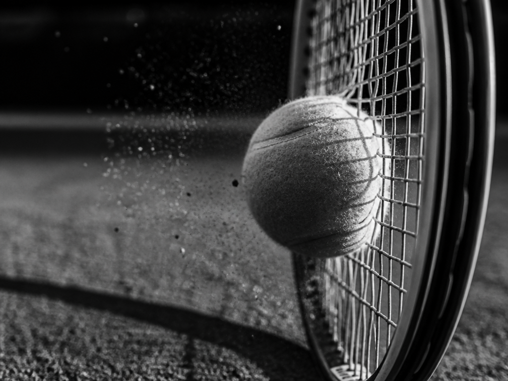 A high-contrast black and white photograph of a tennis ball mid-compression against the string…