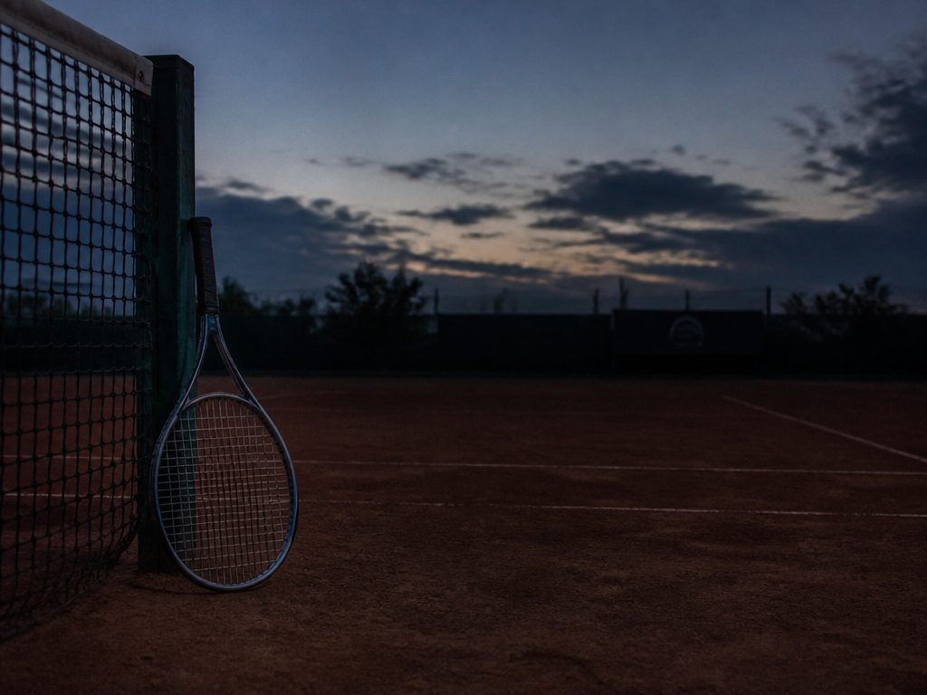 Wide shot of a clay court at dusk, the surface textured and uneven with…