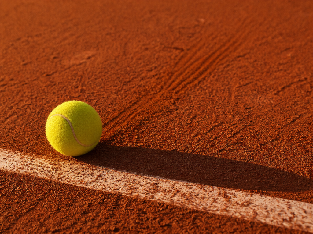 A single tennis ball sits on a red clay court near the baseline, motionless…