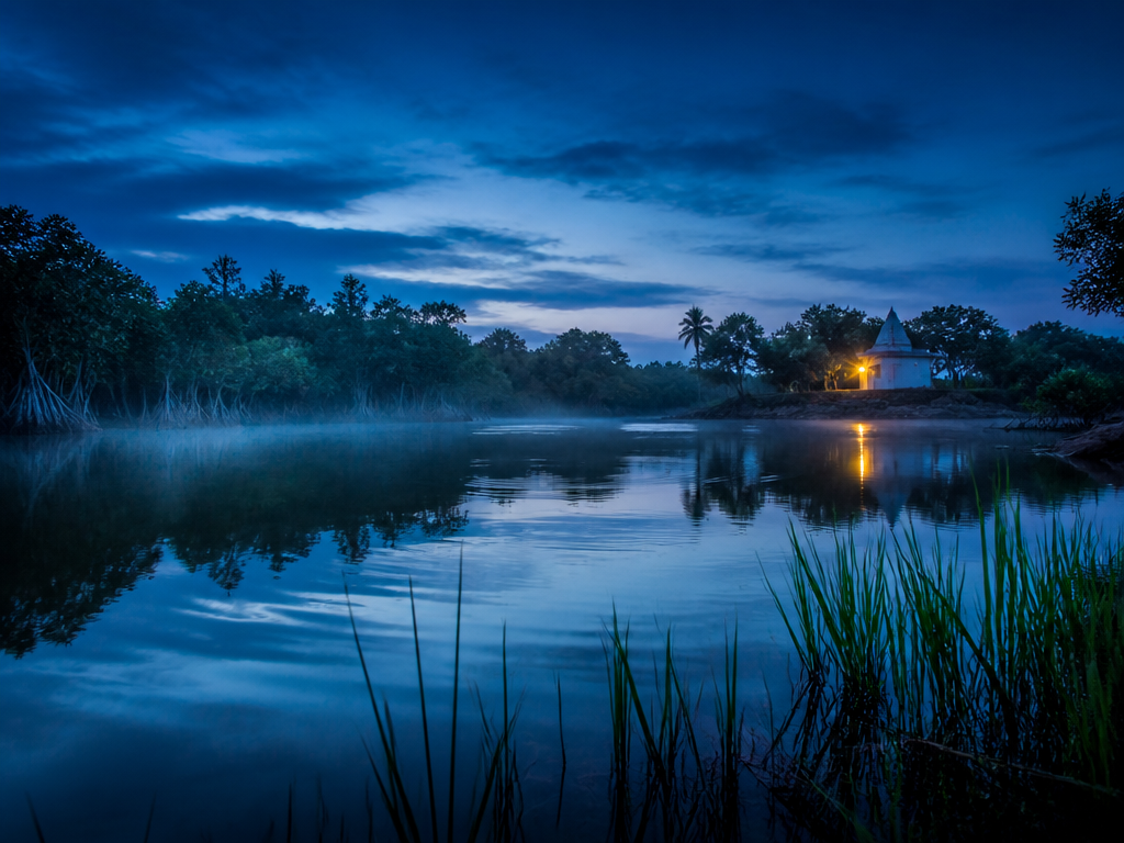 A slow-moving lagoon at blue hour in coastal West Africa, glassy dark water reflecting…