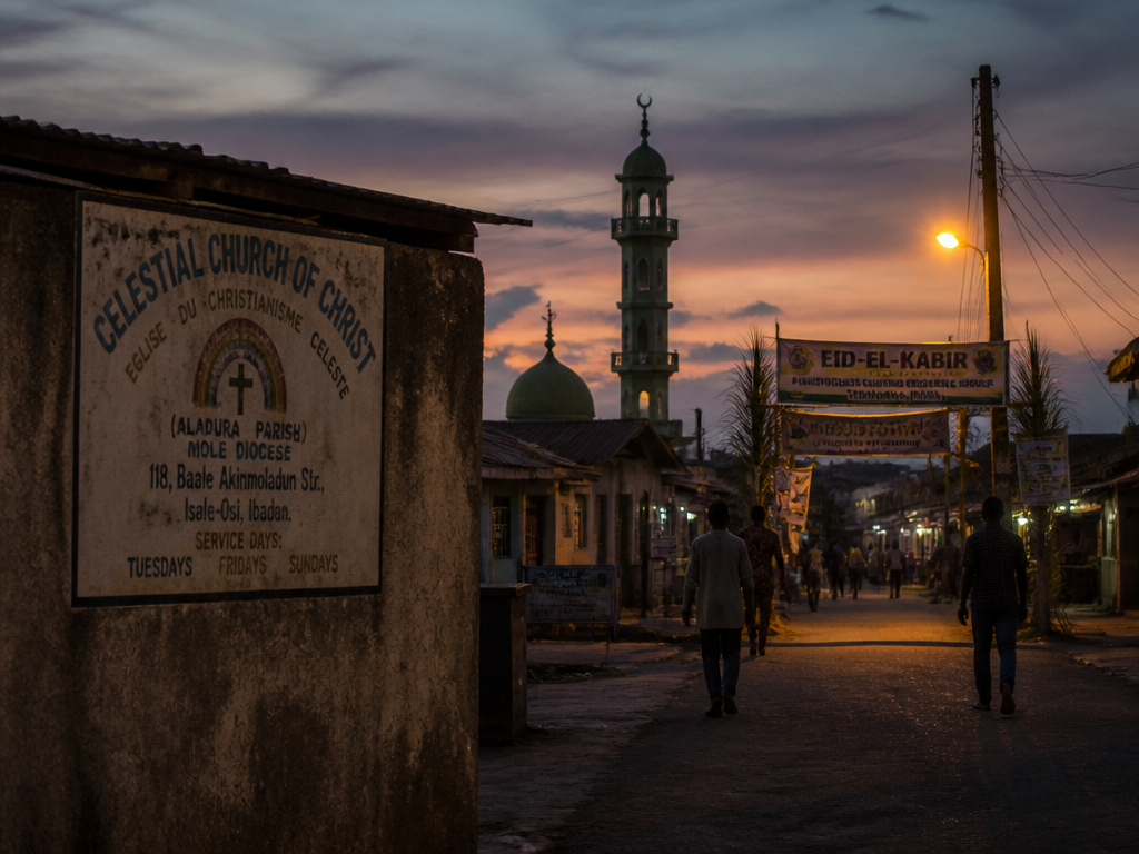 Photorealistic travel-documentary photograph of a Yoruba town street at dusk where two worlds sit…